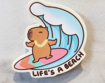 Peut inclure: Un capybara brun portant un collier de fleurs jaunes surfe sur une planche de surf rose et orange avec une vague bleue derrière lui. Le texte "Life's a beach" est écrit sous la planche de surf.