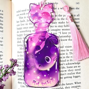 May include: A whimsical bookmark featuring a purple cat inside a potion bottle with a pink tassel. The cat-shaped top has mushroom and cloud designs. The text "Plot twist?" is at the bottom. The bookmark is placed on an open book.