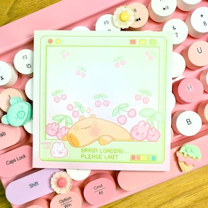 May include: A square, pale green and pink notepad featuring a cartoon capybara, cherries, and the text "BRAIN LOADING... PLEASE WAIT." The notepad is set against a pink keyboard.