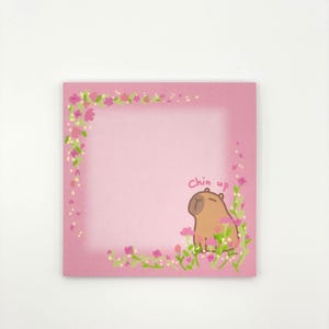 May include: Pink sticky note pad with a floral border and a cartoon capybara with the text "Chin up" in the bottom right corner.