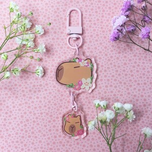May include: A pink and brown capybara keychain with a floral design. The keychain is made of clear acrylic and has a metal clasp.