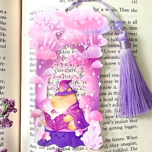 Könnte beinhalten: Ein skurriles Lesezeichen mit einem Cartoon-Charakter in einem Zaubererhut und -umhang, der ein Buch mit dem Titel "Book of Spells" liest. Das Design umfasst rosa und lila Pilze, Wolken und Kristalle, mit einer passenden Quaste.