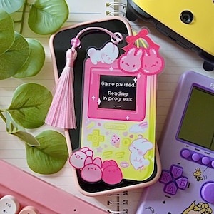 May include: A pink and yellow Game Boy-style phone case with a pink tassel charm. The case has a screen that says "Game paused. Reading in progress." and a cartoon bunny on the bottom.