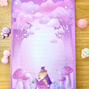 May include: A lined purple notepad with a whimsical forest scene. The illustration features a creature in a wizard hat reading a book, surrounded by mushrooms and crystals. The background has clouds and trees.