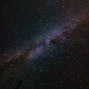 Può includere: Un cielo notturno pieno di stelle, tra cui la Via Lattea, visibile come una fascia di luce attraverso l'immagine. Una sagoma di un palo della linea elettrica si trova in primo piano.