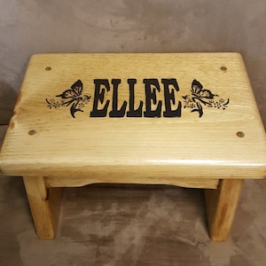 May include: A wooden step stool with a natural finish. The top of the stool is engraved with the name "ELLEE" and two butterflies.