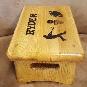 May include: A wooden step stool with a cutout handle. The top of the stool is personalized with the name "RYDER" and images of a football, basketball, and baseball player.