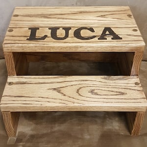 May include: A wooden two-step stool with the name "LUCA" engraved on the top step. The stool is made of light brown wood with a natural wood grain finish.