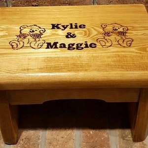 May include: A small, wooden step stool with a light brown finish. The top surface features the names "Kylie & Maggie" and two etched teddy bear designs. The stool has four sturdy legs and is suitable for children.