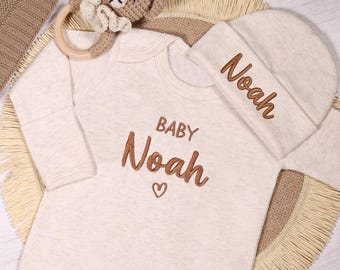 May include: A beige baby onesie with the text "BABY Noah" in brown embroidery. A matching beige knitted hat with the name "Noah" embroidered in white. A brown knitted blanket with the text "Baby Noah" embroidered in white.