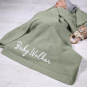 May include: A soft, sage green knitted baby blanket with the name "Baby Walker" embroidered in white.