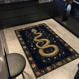 May include: A rectangular area rug featuring a dark navy blue background with a large, central golden snake illustration coiled in a spiral. The rug is bordered by a decorative frame of golden floral and botanical patterns on all four sides.