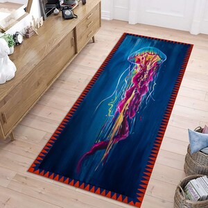 Puede incluir: Una alfombra azul y roja con un diseño de medusa colorido. La medusa está pintada en tonos de rosa, amarillo y azul. La alfombra tiene un borde geométrico con triángulos rojos y azules.