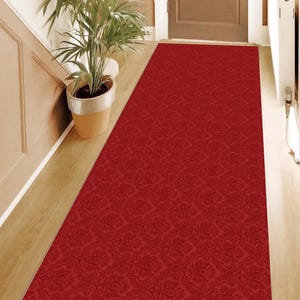 May include: A red runner rug with a damask pattern. The rug is placed in a hallway with a wooden floor and a door to the left.