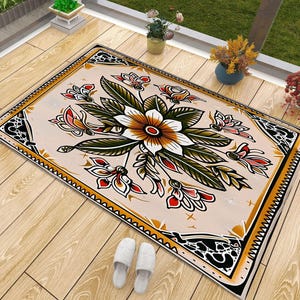 May include: A rectangular area rug featuring a central floral motif with white, orange, and dark green petals surrounded by dark green leaves. The design includes a black and gold decorative border on a light beige background with small star accents.