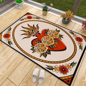 Puede incluir: Una alfombra de color crema con un borde negro y un corazón rojo con flores amarillas. El corazón está rodeado de un marco ovalado negro y dorado. La alfombra tiene un estilo de tatuaje vintage.