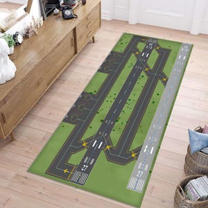 Puede incluir: Una alfombra rectangular con un fondo verde y un diseño detallado de pista de aeropuerto. La alfombra presenta múltiples pistas, calles de rodaje y marcas, incluido el número "27". El diseño incorpora elementos grises, negros, blancos y amarillos, creando un diseño realista del aeropuerto.