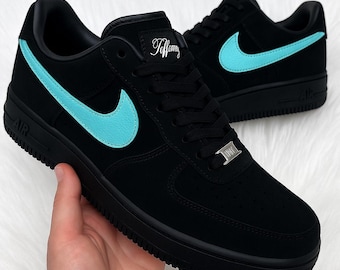 May include: Black Air Force 1 sneakers with a turquoise Nike swoosh. The sneakers have black laces, soles, and a small silver tag. The word "Tiffany" is written in cursive on the tongue.