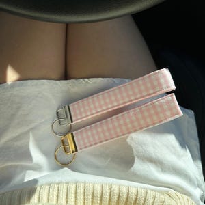 May include: Two key fobs with pink and white gingham fabric straps. One has a silver-colored metal ring and hardware, and the other has gold-colored hardware. The key fobs are lying on a white surface.