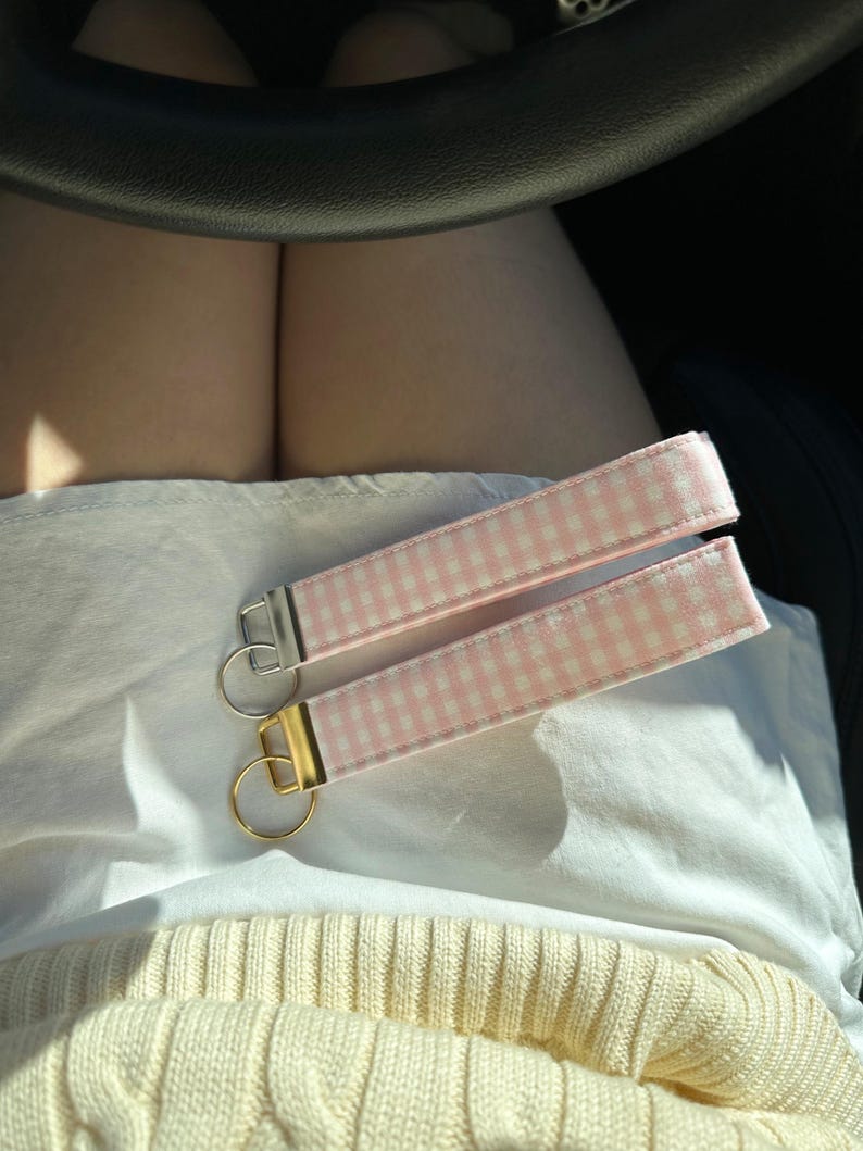 May include: Two key fobs with pink and white gingham fabric straps. One has a silver-colored metal ring and hardware, and the other has gold-colored hardware. The key fobs are lying on a white surface.