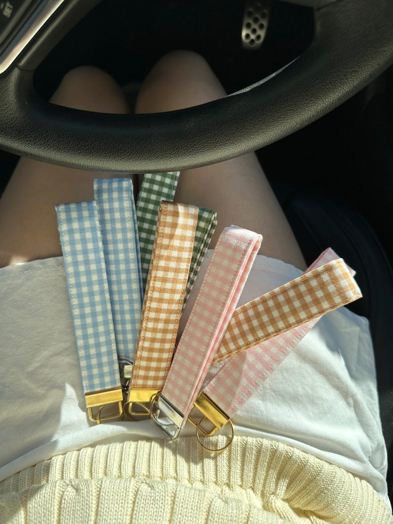 May include: A collection of colorful gingham key fobs in various shades of blue, green, gold, and pink. Each fob has a gold or silver-colored metal clasp. The key fobs are displayed on a person's lap.