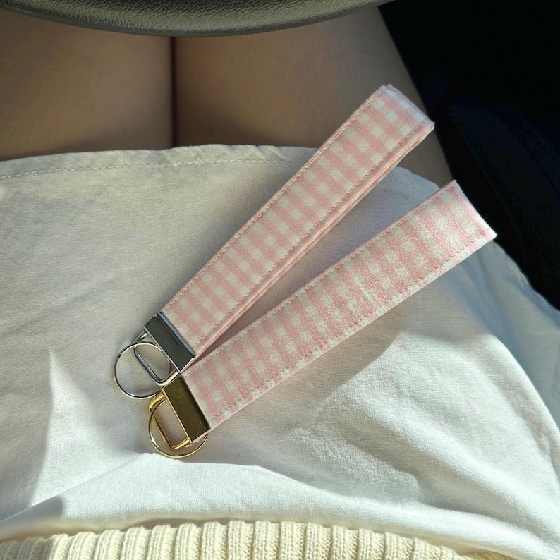 May include: Two pink and white gingham key fobs with metal hardware. One fob has a silver-colored ring and hardware, while the other has gold-colored hardware. The key fobs are lying on a white surface.