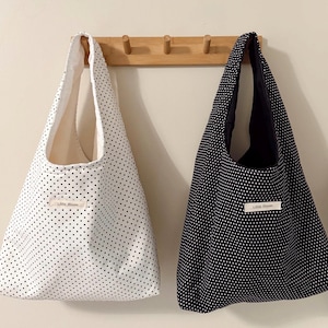 May include: Two tote bags hanging on a wooden rack. One bag is white with black polka dots, the other is black with white polka dots. Each bag has a label that reads "Little Bloom".