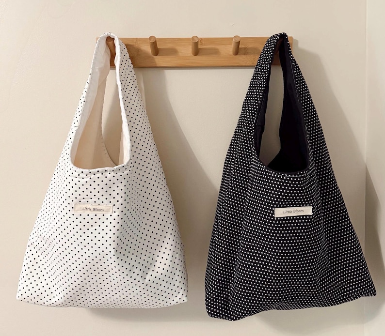 May include: Two tote bags hanging on a wooden rack. One bag is white with black polka dots, the other is black with white polka dots. Each bag has a label that reads "Little Bloom".
