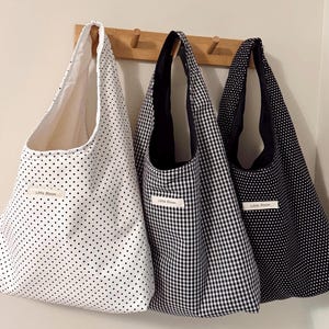 May include: Three tote bags hanging on a wooden rack. The bags feature different patterns: white with black polka dots, black and white gingham, and navy with white polka dots. Each bag has a label that reads "Little Bloom".