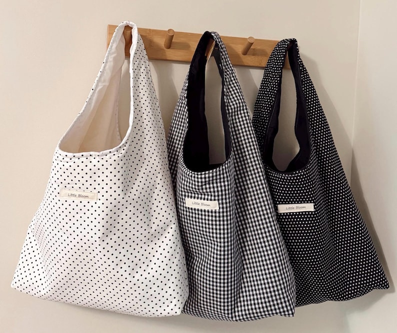 May include: Three tote bags hanging on a wooden rack. The bags feature different patterns: white with black polka dots, black and white gingham, and navy with white polka dots. Each bag has a label that reads "Little Bloom".