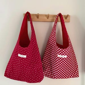Op de afbeelding: Twee rode draagtassen met witte stippen hangen aan een houten rek. De tassen hebben een eenvoudig ontwerp met lange hengsels en een contrasterende rode binnenkant. Elke tas heeft een klein wit label met de tekst "Little Bloom".