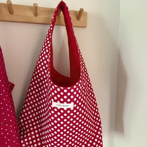 Op de afbeelding: Een rode draagtas met witte stippen hangt aan een houten rek. De binnenkant van de tas is effen rood en een klein label leest "Little Bloom". De tas is gemaakt van stof en heeft een eenvoudig, open ontwerp.