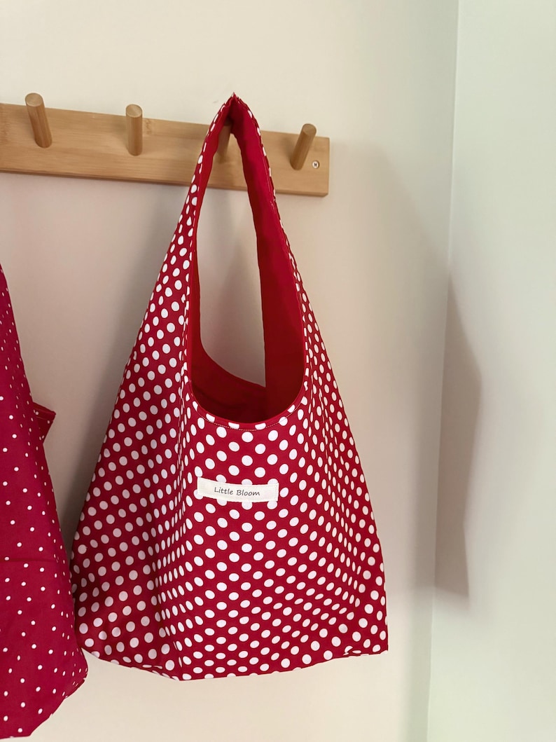 Op de afbeelding: Een rode draagtas met witte stippen hangt aan een houten rek. De binnenkant van de tas is effen rood en een klein label leest "Little Bloom". De tas is gemaakt van stof en heeft een eenvoudig, open ontwerp.