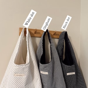 May include: Three tote bags hang on a wooden rack. The bags are white with black dots, black and white gingham, and black with white dots. Each bag has a label that reads "Little Bloom". The tags above the bags read "White dot", "Gingham", and "Black dot".