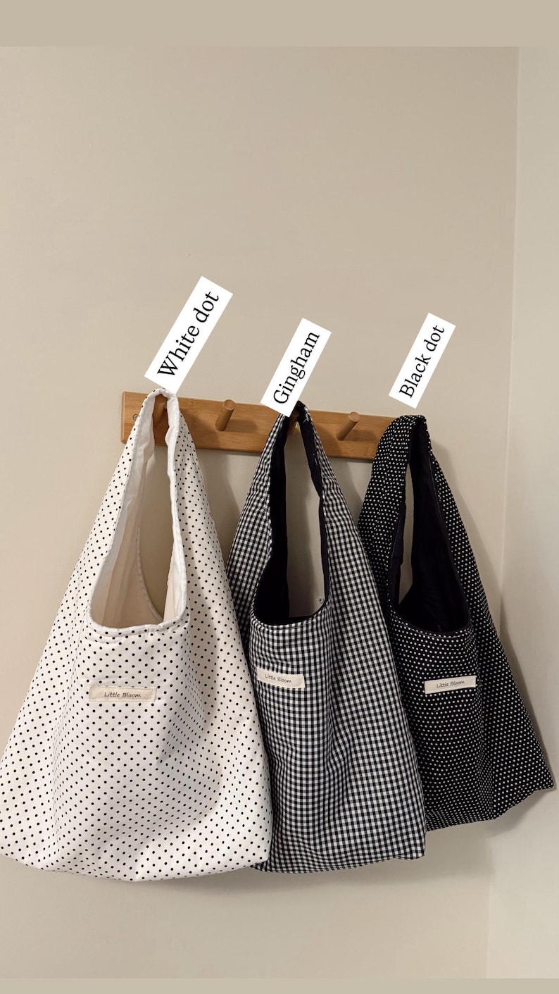 May include: Three tote bags hang on a wooden rack. The bags are white with black dots, black and white gingham, and black with white dots. Each bag has a label that reads "Little Bloom". The tags above the bags read "White dot", "Gingham", and "Black dot".