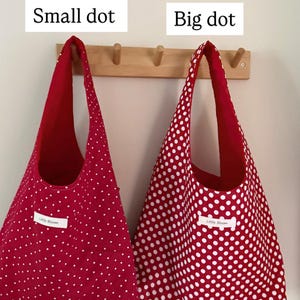 Op de afbeelding: Twee rode draagtassen met witte stippen hangen aan een houten rek. E&eacute;n tas heeft kleine stippen, de andere heeft grote stippen. De tassen zijn gelabeld met "Small dot" en "Big dot". Elke tas heeft een wit label met "Little Bloom".