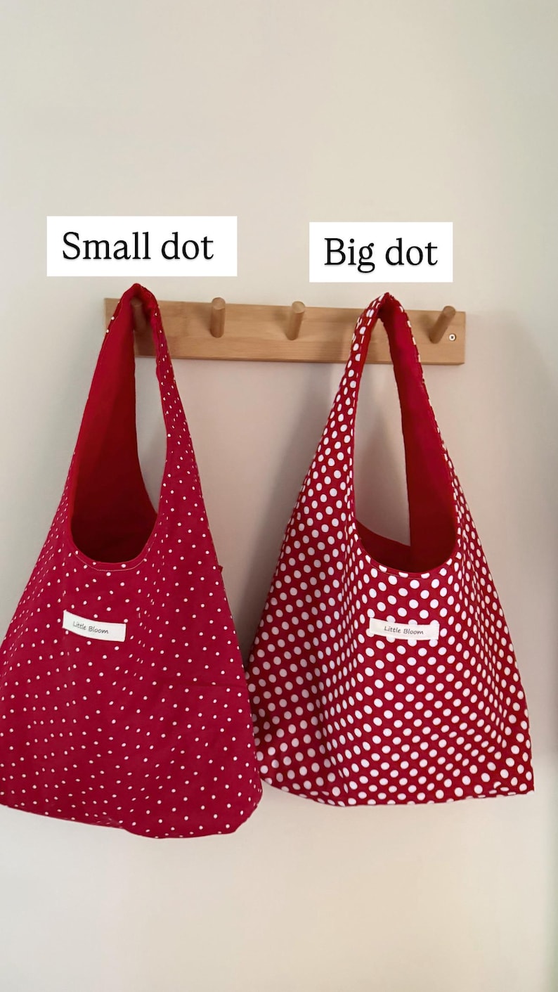 Op de afbeelding: Twee rode draagtassen met witte stippen hangen aan een houten rek. E&eacute;n tas heeft kleine stippen, de andere heeft grote stippen. De tassen zijn gelabeld met "Small dot" en "Big dot". Elke tas heeft een wit label met "Little Bloom".