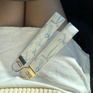May include: Two white fabric keychains with gold key rings. The keychains feature a blue and white pattern of butterflies.
