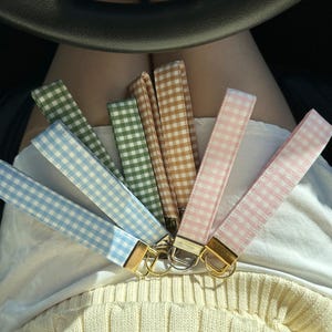 May include: A set of six colorful gingham keychains with gold key rings. The keychains are in shades of blue, green, brown, pink, and light pink.