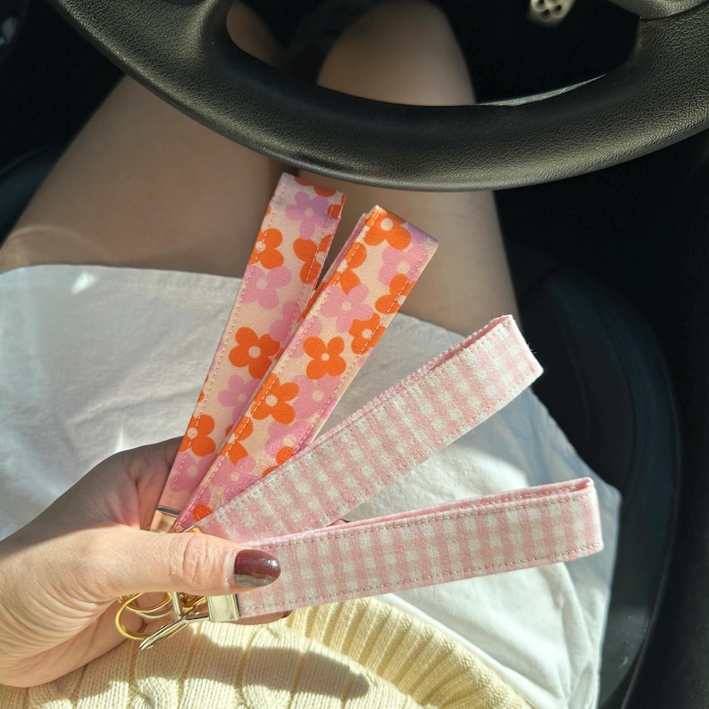 May include: Three fabric keychains in the image. Two are patterned with pink and orange flowers, and the third is a pink and white gingham pattern. The keychains have gold-colored hardware.