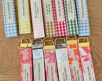 May include: A collection of fabric keychains with various patterns and text. The keychains feature gingham, floral, and patterned designs. Each keychain has a metal ring and a label with phrases like "You are doing great" and "God has my back."