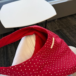 Op de afbeelding: Een rode schoudertas met witte stippen. De tas heeft een gebogen vorm en een lange riem. De binnenkant is effen rood. Een klein wit label met tekst is op de tas genaaid.