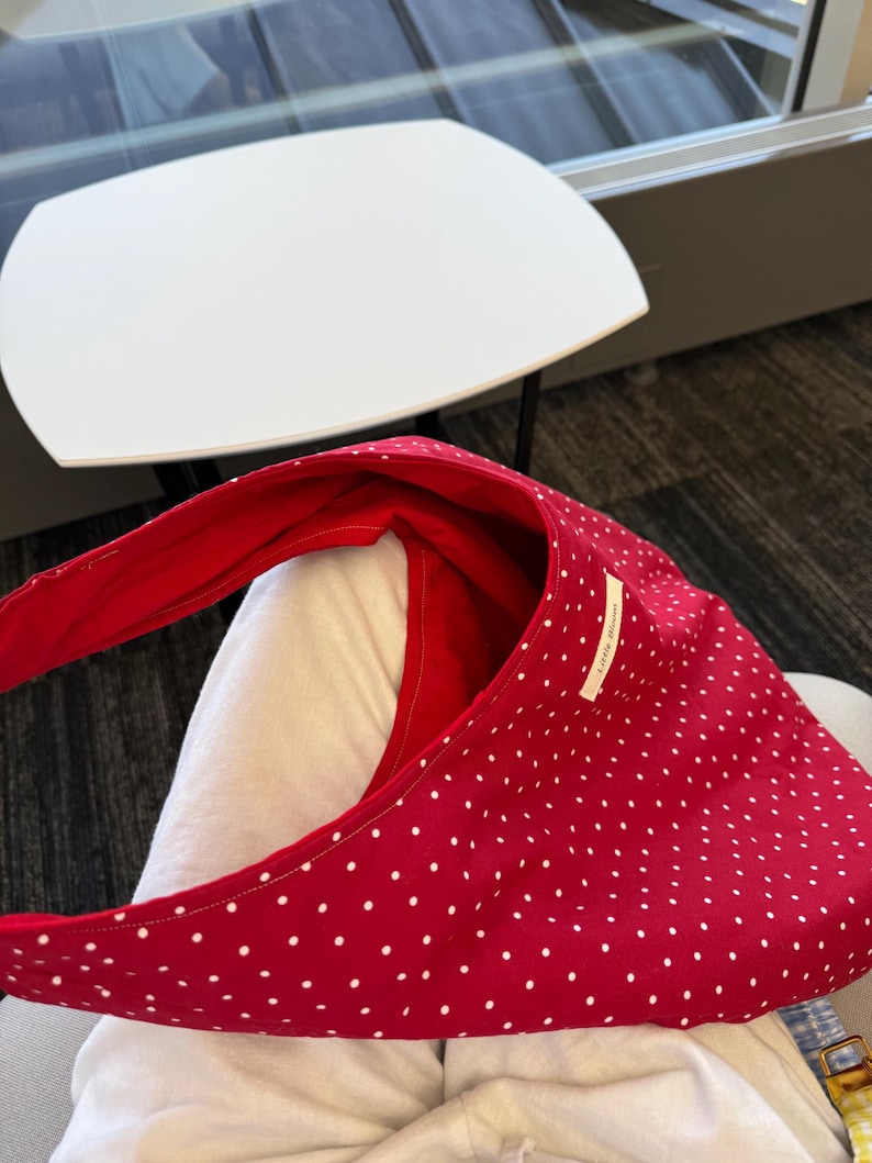 Op de afbeelding: Een rode schoudertas met witte stippen. De tas heeft een gebogen vorm en een lange riem. De binnenkant is effen rood. Een klein wit label met tekst is op de tas genaaid.