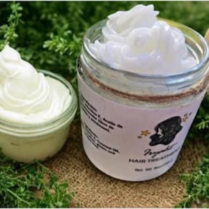 May include: Two glass jars filled with white cream, one small and one large, sit on a bed of green foliage. The larger jar has a label with text and a silhouette. A wooden spoon rests on the rim of the larger jar.