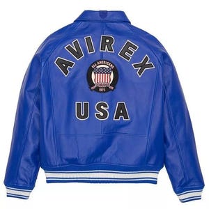May include: A vibrant blue leather jacket with the word "AVIREX" arched across the back in white block letters. Below the name is a circular patch with the words "ALL AMERICANS ESTABLISHED 1975" and a USA flag design. The word "USA" is printed below the patch.