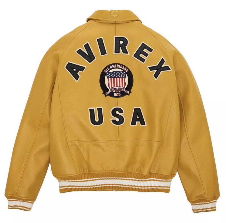 May include: Mustard yellow leather jacket with the word "AVIREX" across the back in black lettering. Below the word "AVIREX" is a patch with the words "ALL AMERICANS ESTABLISHED 1975" and a graphic of the American flag. The word "USA" is below the patch.