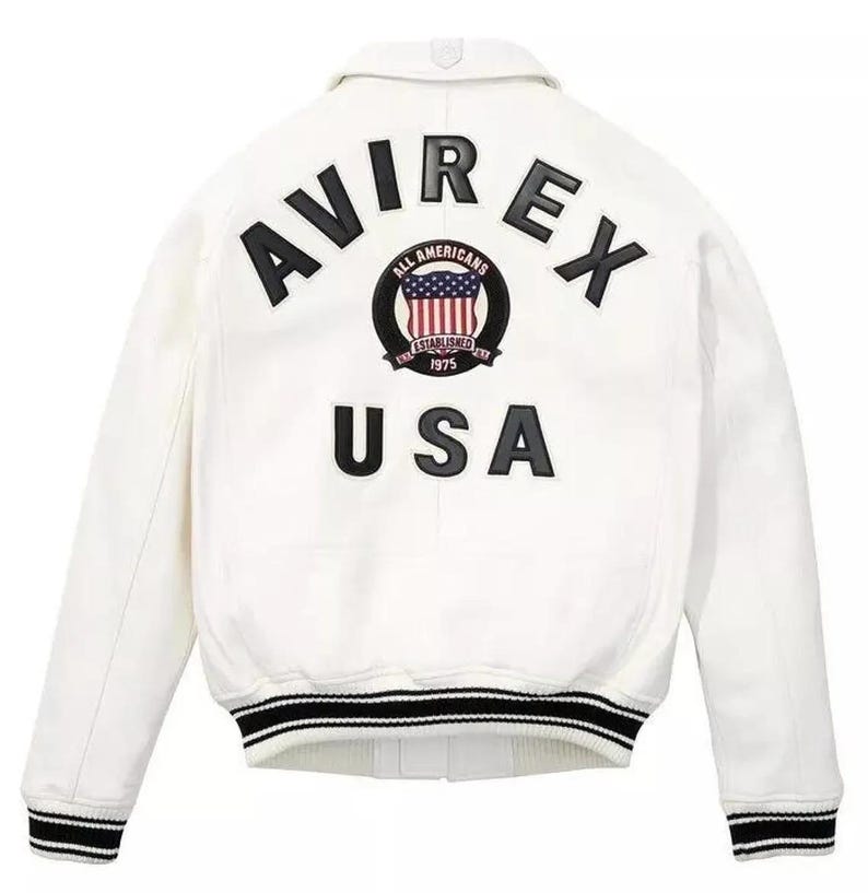 May include: White jacket with black and white striped trim on the cuffs and hem. The back features the text "AVIREX USA" in black, with a circular emblem of the American flag and the words "ALL AMERICANS ESTABLISHED 1975".