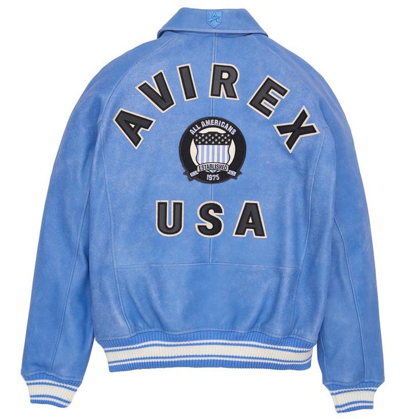 May include: Light blue leather jacket with the word "AVIREX" arched across the back, above a circular patch that reads "ALL AMERICANS ESTABLISHED 1975". Below is the word "USA". The jacket has white and blue striped cuffs and a collar.