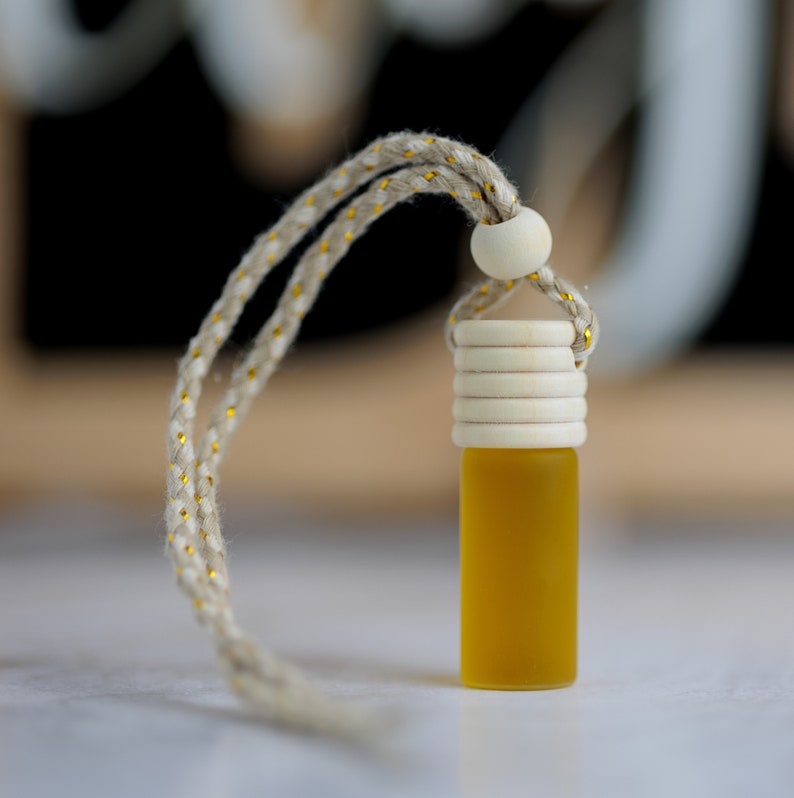 May include: A small, yellow glass bottle with a wooden cap and a beige rope. The rope has gold accents and is attached to a wooden bead. The bottle is filled with a yellow liquid. The background is blurred.