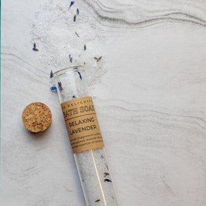 Puede incluir: Un tubo de vidrio lleno de sales de ba&ntilde;o blancas y flores de lavanda. La etiqueta dice "E DELIGHTFUL BATH SOAK RELAXING LAVENDER salt (magnesium sulfate), dried cyanus, lavender flowers, lavender essential oil (lavender"